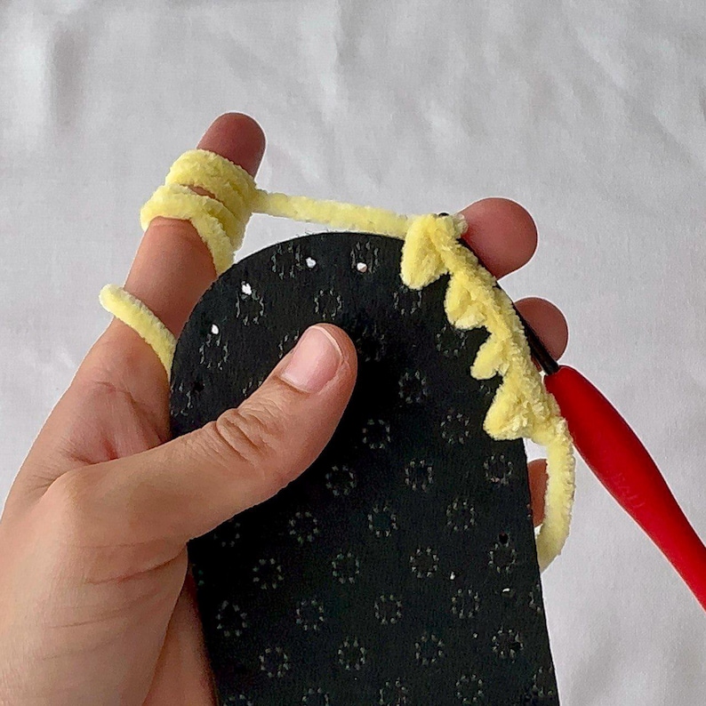 以下が含まれることがあります： 黄色の糸でかぎ針編みされた黒いスリッパの底のクローズアップ。赤いかぎ針編みフックが見え、糸が指に巻き付いています。クラフトプロジェクトの進行状況を示しています。