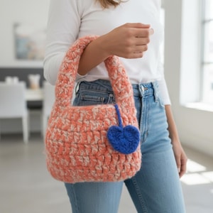 Könnte beinhalten: Eine gehäkelte Handtasche in Pfirsich und Creme mit einem herzförmigen Anhänger in Blau. Die Tasche hat eine runde Form mit zwei Henkeln. Der Anhänger ist mit einer blauen Schnur an der Tasche befestigt.