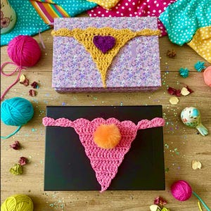 以下が含まれることがあります： 手作りのかぎ針編みのTバック2つ。マスタードイエローに紫色のハート、ピンクにオレンジ色のポンポン。装飾的な箱の上に置かれ、糸やカラフルな生地に囲まれています。