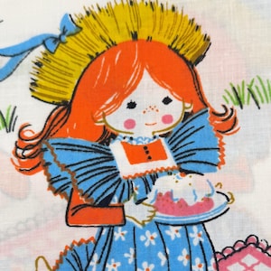Op de afbeelding: Een witte stof met een kleurrijke illustratie van een persoon met oranje haar die een strooien hoed en een blauwe bloemenjurk draagt. De persoon houdt een bord met een taart vast. Onderaan ligt een roze geblokt picknickkleed met een theepot.