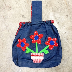 以下が含まれることがあります： デニムのトートバッグ、花の模様。 赤い花のアップリケが3つ、青い中心と緑の茎があり、ギンガムの鉢に入っています。 ハンドルはデニムで、赤いトリムが付いています。