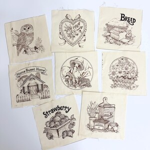May include: Seven square off-white fabric patches featuring black line art illustrations. Designs include an owl, a heart with roses, a loaf of bread with the text BREAD, a house with Home Sweet Home, mushrooms, a flower basket, strawberries, and kitchen tools.