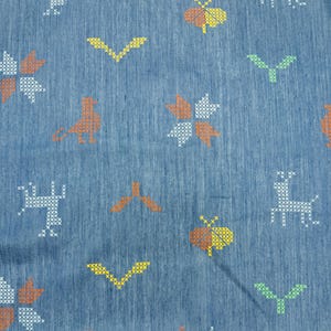 May include: Close-up of a blue fabric with a cross-stitch pattern. The pattern includes various shapes such as birds, flowers, and insects in orange, yellow, white, and green. The fabric has a textured appearance.