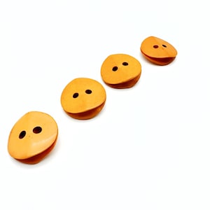 Puede incluir: Cuatro botones de color amarillo anaranjado con dos agujeros cada uno, dispuestos en diagonal sobre un fondo blanco. Los botones tienen una forma redondeada y ligeramente curvada, lo que sugiere un diseño único para coser o manualidades.