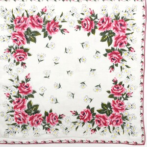 May include: A square white handkerchief featuring a floral pattern with clusters of pink roses and green leaves in the corners. The center and edges are decorated with small white daisy-like flowers and a thin pink border along the perimeter.