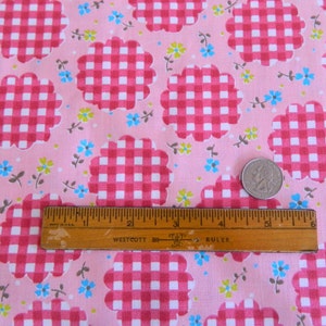 May include: Pink fabric with a repeating pattern of white and pink gingham squares in a scalloped flower shape. Scattered throughout the fabric are small blue, green, and brown flowers. A ruler and a coin are included in the image for scale.