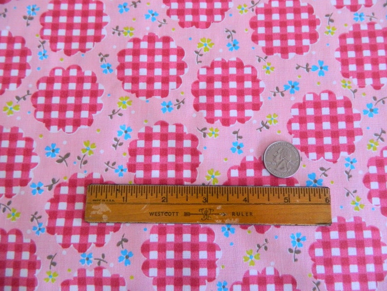 May include: Pink fabric with a repeating pattern of white and pink gingham squares in a scalloped flower shape. Scattered throughout the fabric are small blue, green, and brown flowers. A ruler and a coin are included in the image for scale.