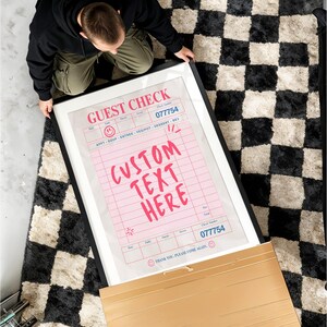 May include: A framed print featuring a pink guest check design with the text "CUSTOM TEXT HERE". The print has a black frame and is on a black and white checkered rug. The check includes the check number 077754.
