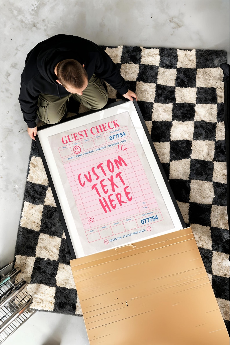May include: A framed print featuring a pink guest check design with the text "CUSTOM TEXT HERE". The print has a black frame and is on a black and white checkered rug. The check includes the check number 077754.
