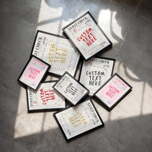 May include: A collection of framed prints featuring a guest check design. Each print has the words "GUEST CHECK" and "CUSTOM TEXT HERE" in various colors, including red, pink, and gold. The frames are black.