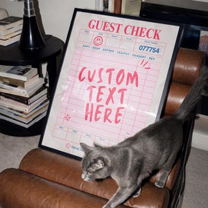 May include: A framed pink and white "Guest Check" print with the text "CUSTOM TEXT HERE". A gray cat walks across a brown leather chair. A black lamp and books are in the background.