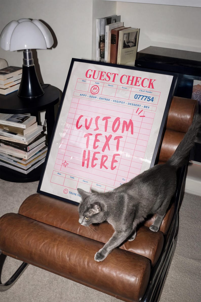 May include: A framed pink and white "Guest Check" print with the text "CUSTOM TEXT HERE". A gray cat walks across a brown leather chair. A black lamp and books are in the background.