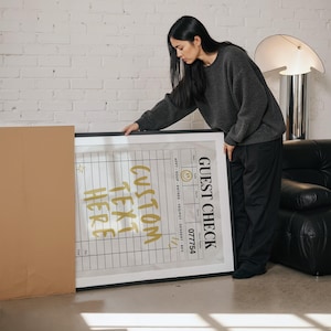 May include: A framed print with the words "CUSTOM TEXT HERE" in gold lettering. The print also includes a "GUEST CHECK" section with a grid and the number "077754". The frame is black.