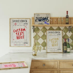 May include: A kitchen scene featuring framed "Guest Check" prints with "Custom Text Here" in red and yellow. Wine bottles, champagne, and glasses are arranged on a countertop and shelf. A wooden tray with a pink guest check is in the foreground.