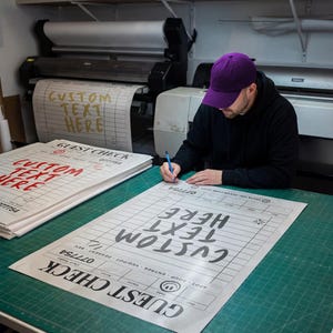May include: A person wearing a purple cap is writing on a large printed guest check with the words "CUSTOM TEXT HERE". The check is on a green cutting mat. Other printed guest checks are stacked nearby.