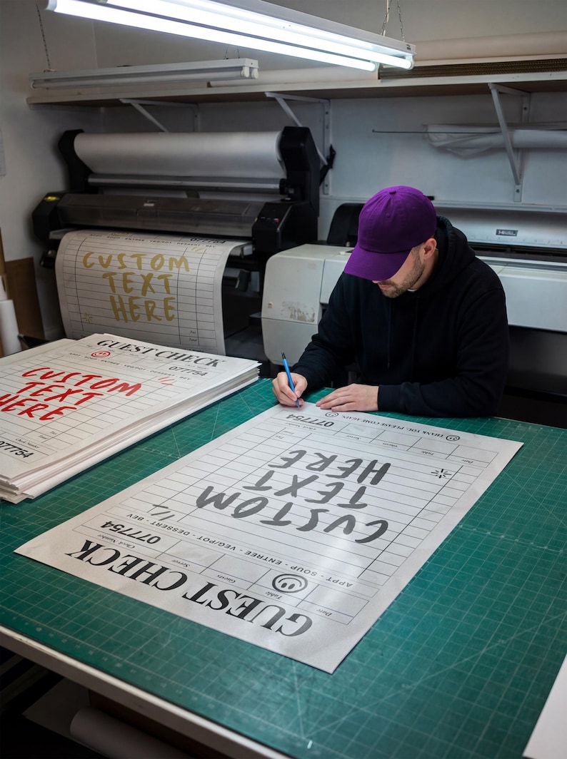 May include: A person wearing a purple cap is writing on a large printed guest check with the words "CUSTOM TEXT HERE". The check is on a green cutting mat. Other printed guest checks are stacked nearby.