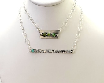 May include: A silver necklace with two pendants. The top pendant is a rectangular bar with three green stones. The bottom pendant is a long bar with a floral design and a turquoise stone.