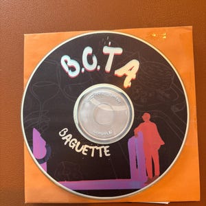 Könnte beinhalten: Eine CD mit schwarzem Hintergrund mit dem Text "B.C.T.A." in Weiß und Rosa und "BAGUETTE" in Weiß. Die Disc hat eine farbenfrohe Illustration einer Person und einer violetten Struktur. Die CD befindet sich in einer orangefarbenen Papierhülle.