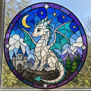 Könnte beinhalten: Ein runder Buntglas-Sonnenfänger mit einem weißen Drachen mit türkisfarbenen Flügeln auf einem Felsen. Der Hintergrund zeigt einen Nachthimmel mit gelbem Halbmond, Sternen, einer grauen Steinburg, violetten Bergen und grünen Tannenbäumen.