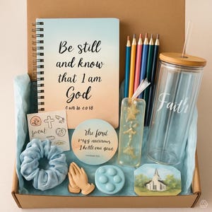 May include: A gift box containing stationery and accessories. It features a spiral-bound notebook with the text "Be still and know that I am God", pencils, a glass tumbler with "Faith", a scrunchie, and a bookmark.