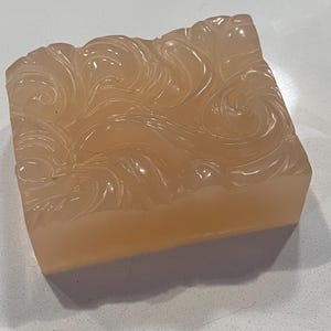 May include: A rectangular bar of peach-colored soap with a carved, swirling design on the top surface. The soap has a translucent quality, and the carved pattern creates shadows. The bottom edge of the soap is a solid peach color.