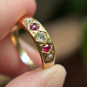 Könnte beinhalten: Ein Ring aus poliertem Gelbgold mit einem abwechselnden Muster aus zwei runden roten Edelsteinen und drei klaren, facettierten weißen Edelsteinen. Jeder Stein ist in einer sternförmigen Gravur auf der Oberfläche des Goldbandes gefasst.