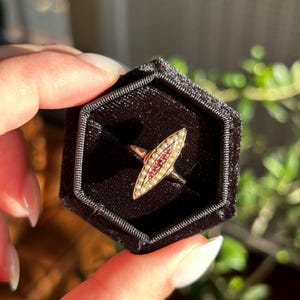 Pode incluir: Um anel de ouro com um design em forma de marquesa, cravejado com pequenas pérolas e pedras preciosas vermelhas. O anel é exibido em uma caixa hexagonal de veludo preto. A banda do anel é prateada.