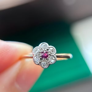 Pode incluir: Um anel com uma pedra vermelha central de corte redondo rodeada por seis pequenas pedras claras de corte redondo dispostas num padrão floral. A cravação é de metal prateado e o aro é de metal dourado fino e polido.