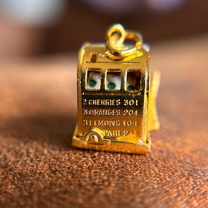 May include: A small, gold-colored slot machine charm with a loop at the top. The charm has three windows displaying green and white symbols. Text on the front reads "2 Cherries 30:1", "3 Oranges 20:1", "3 Lemons 10:1", and "Pair 2:1".