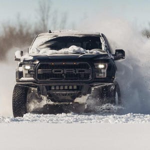 May include: A black Ford Raptor truck driving through deep snow, creating a large cloud of snow. The truck has a black grill and headlights. The scene is set in a snowy field with trees in the background.