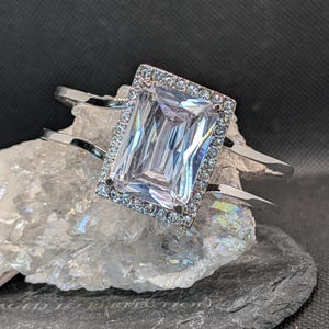 May include: A silver-toned ring with a large, rectangular, clear gemstone. The gemstone is surrounded by smaller, clear stones. The ring is displayed on a white, crystalline surface against a dark background.