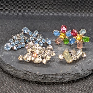 May include: A collection of four vintage-style brooches featuring various designs. One brooch is light blue, one is clear, one is multi-colored with pink, yellow, green, and blue stones, and one is a small rectangular clear stone piece.