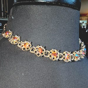 May include: A silver-toned metal choker necklace featuring a series of connected floral-shaped links, each set with a round, iridescent orange and gold faceted stone that reflects multiple colors in the light.