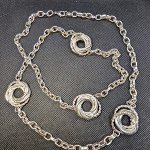 May include: Silver-toned necklace featuring a chain design with five decorative circular elements. Each circular element has a twisted rope-like design in the center. The necklace is displayed on a black surface.