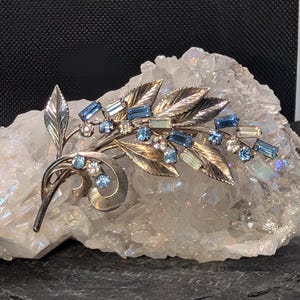 May include: A silver-toned metal brooch shaped like a leafy branch, featuring a collection of light blue and clear faceted rhinestones set along the stems and leaves. The design includes curved metal elements and detailed leaf textures.