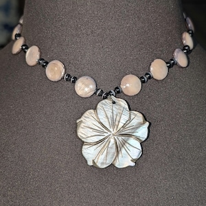 May include: A necklace featuring a large, carved, iridescent shell pendant shaped like a five-petaled flower. The chain consists of alternating round, light-toned, polished shell beads and small, dark, banded spherical beads strung together.