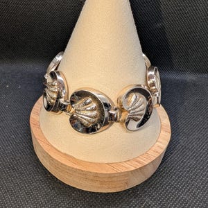 May include: A silver-toned bracelet with a seashell design. The bracelet is composed of oval links, each featuring a detailed seashell motif. The bracelet is displayed on a beige, cone-shaped stand, set against a dark background.