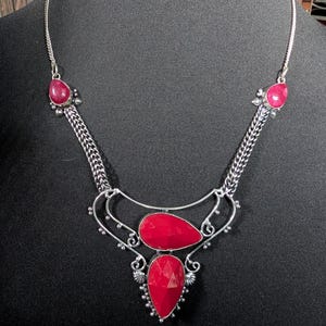 May include: A silver-toned necklace featuring four faceted, teardrop-shaped red stones. The central pendant consists of two stacked red stones framed by ornate silver wirework, connected to two smaller red stones by textured silver chain links.