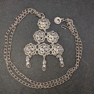 May include: A silver-toned metal necklace featuring a multi-tiered pendant composed of six interconnected floral filigree motifs. Three small floral drops hang from the bottom of the pendant. The chain is a simple link style with a hook clasp.