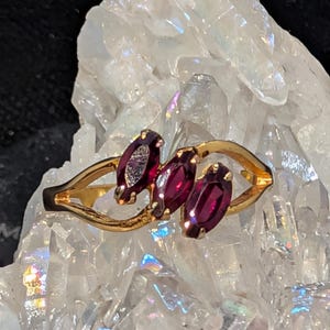 May include: A gold ring featuring three oval, deep red gemstones. The ring's band has a delicate, flowing design. The gemstones are set in a row, creating a striking visual contrast against the gold. The ring is displayed on a clear, crystalline surface.
