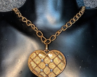 May include: A gold-toned necklace featuring a large heart-shaped pendant with a textured, quilted grid pattern. The pendant is accented with five small, clear, round-cut stones embedded within the grid. The chain consists of thick, oval-shaped links.