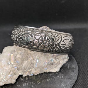 May include: A silver-toned bangle bracelet with intricate floral carvings. The bracelet is wide and features a repeating pattern of flowers and leaves. The bracelet is displayed on a white crystal formation and a black stone with the text "VINTAGE AGED TO PERFECTION".
