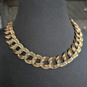 May include: A gold-toned metal chain necklace featuring a series of interlocking, textured circular links. The necklace has a substantial, heavy appearance with a brushed finish that highlights the layered, ribbed detail on each individual link.