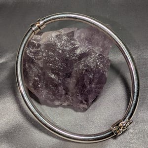 May include: A polished silver-tone hinged bangle bracelet with a smooth, rounded profile and a secure box clasp mechanism, encircling a rough, jagged piece of translucent purple amethyst crystal with natural crystalline textures and facets.
