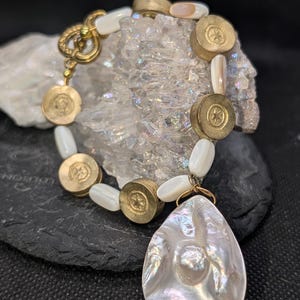 May include: A handcrafted bracelet featuring a teardrop-shaped, iridescent pendant. The bracelet includes gold-toned metal accents, round brass beads with starburst designs, and white, oval-shaped beads. The clasp is a decorative gold-toned hook.