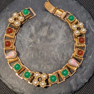 May include: A gold-tone link bracelet featuring alternating square settings with green cabochons, reddish-brown cabochons, iridescent teardrop-shaped stones, and clusters of four small white pearls, secured with a fold-over clasp closure.