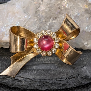 May include: A gold-toned metal brooch shaped like a bow, featuring a central circular cluster of clear rhinestones surrounding a smooth, round, deep pink cabochon stone. The bow has a polished metallic finish with curved, ribbon-like loops.