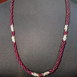 May include: A necklace featuring a twisted design of deep burgundy beads interspersed with small, irregular white pearls. The strands are woven together in a spiral pattern, creating a textured look with consistent spacing between the pearl accents.