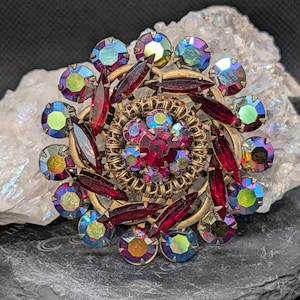 May include: A circular brooch featuring a central red faceted stone surrounded by smaller iridescent blue and green stones, accented with deep red marquise-shaped crystals set in a textured gold-tone metal frame with a layered floral design.