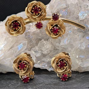 May include: A gold-tone metal brooch and matching clip-on earrings set. Each piece features textured rose-shaped designs with deep red faceted stones set in the center and as small accents. The brooch has a curved bar connecting three roses.